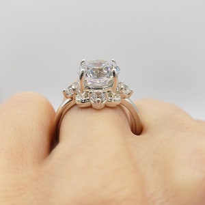 Puede incluir: Un anillo de plata con una piedra central grande, redonda y transparente, rodeada de piedras m&aacute;s peque&ntilde;as y transparentes. El anillo est&aacute; en un dedo.
