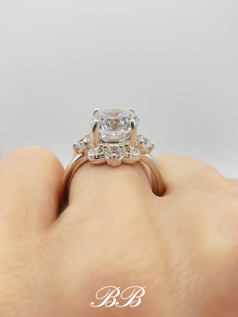 Puede incluir: Un anillo de plata con una piedra central grande, redonda y transparente, rodeada de piedras m&aacute;s peque&ntilde;as y transparentes. El anillo est&aacute; en un dedo.
