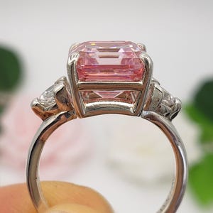 Puede incluir: Un anillo plateado con una gran gema rosa rectangular y dos piedras claras m&aacute;s peque&ntilde;as. El anillo tiene una banda pulida, con las iniciales "BB" visibles. Una joya elegante y llamativa.