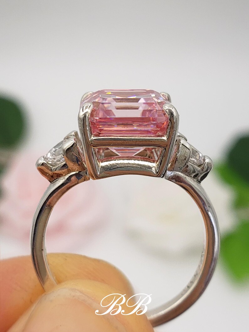 Puede incluir: Un anillo plateado con una gran gema rosa rectangular y dos piedras claras m&aacute;s peque&ntilde;as. El anillo tiene una banda pulida, con las iniciales "BB" visibles. Una joya elegante y llamativa.