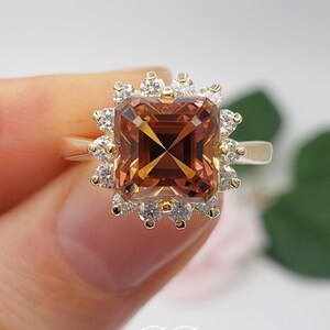 Pode incluir: Um anel de ouro com uma grande gema marrom de forma quadrada, cercada por um halo de gemas menores e transparentes.