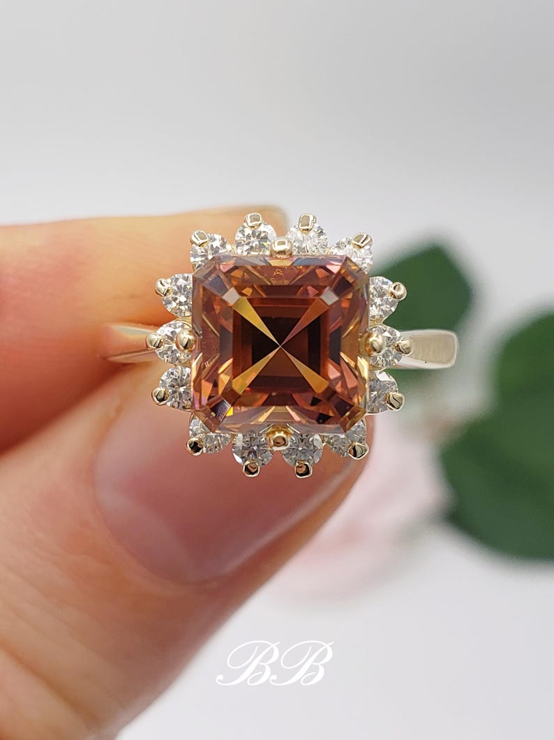 Pode incluir: Um anel de ouro com uma grande gema marrom de forma quadrada, cercada por um halo de gemas menores e transparentes.