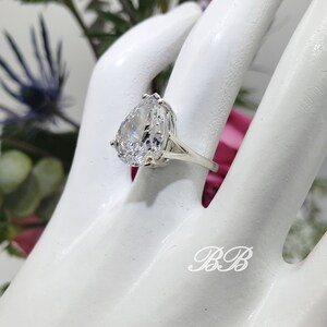 May include: A silver ring with a large pear-shaped clear stone set in a prong setting. The ring is on a white mannequin hand.