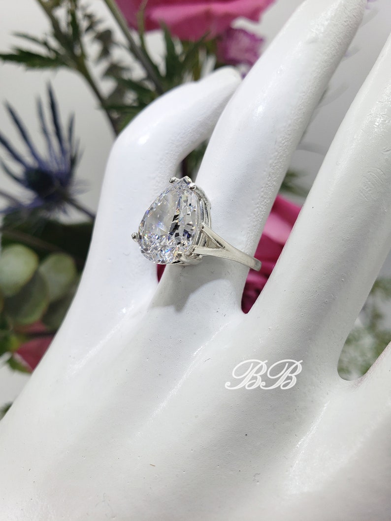 May include: A silver ring with a large pear-shaped clear stone set in a prong setting. The ring is on a white mannequin hand.