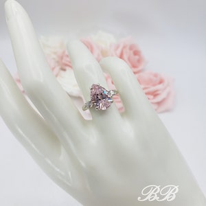 Puede incluir: Un anillo de piedra preciosa rosa en forma de pera con piedras preciosas transparentes m&aacute;s peque&ntilde;as en la banda. El anillo est&aacute; en una mano de maniqu&iacute; blanca.