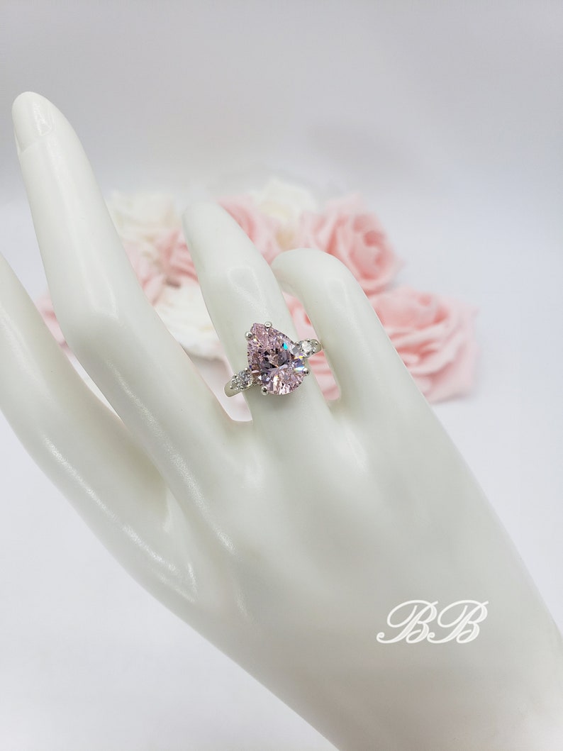 Puede incluir: Un anillo de piedra preciosa rosa en forma de pera con piedras preciosas transparentes m&aacute;s peque&ntilde;as en la banda. El anillo est&aacute; en una mano de maniqu&iacute; blanca.