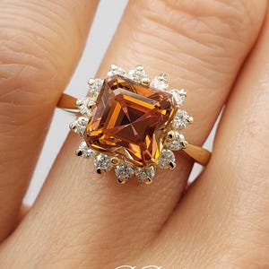 Pode incluir: Um anel de ouro com uma grande gema quadrada, de cor laranja e marrom, cercada por gemas menores, transparentes.