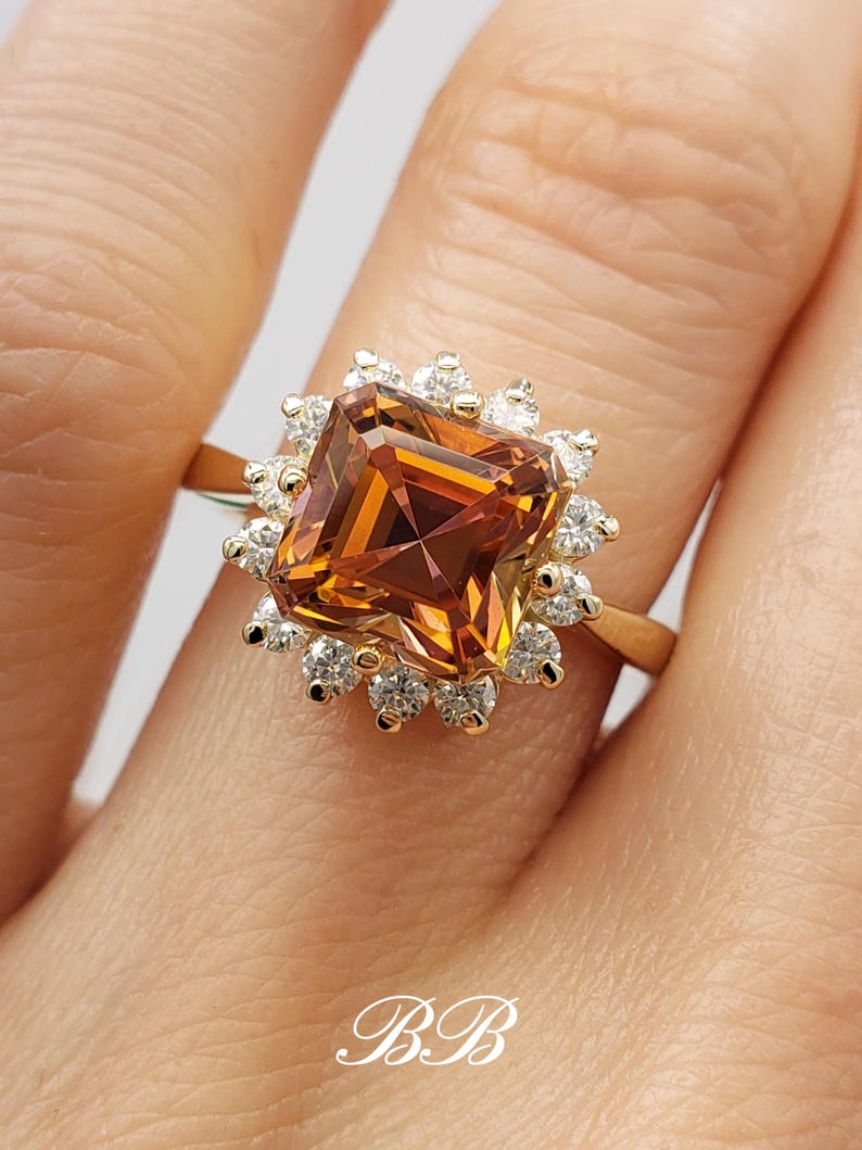 Pode incluir: Um anel de ouro com uma grande gema quadrada, de cor laranja e marrom, cercada por gemas menores, transparentes.