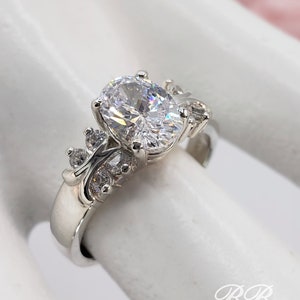May include: A silver ring with a large oval-shaped clear stone surrounded by smaller clear stones. The ring is set on a white background.