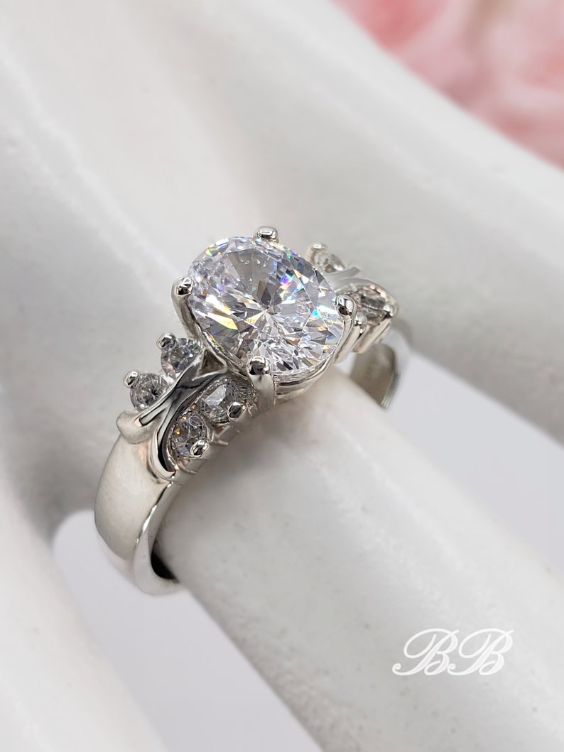 May include: A silver ring with a large oval-shaped clear stone surrounded by smaller clear stones. The ring is set on a white background.