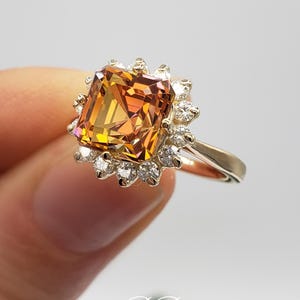 Pode incluir: Um anel de ouro com uma grande gema quadrada, de cor laranja e marrom, cercada por um halo de gemas brancas menores.