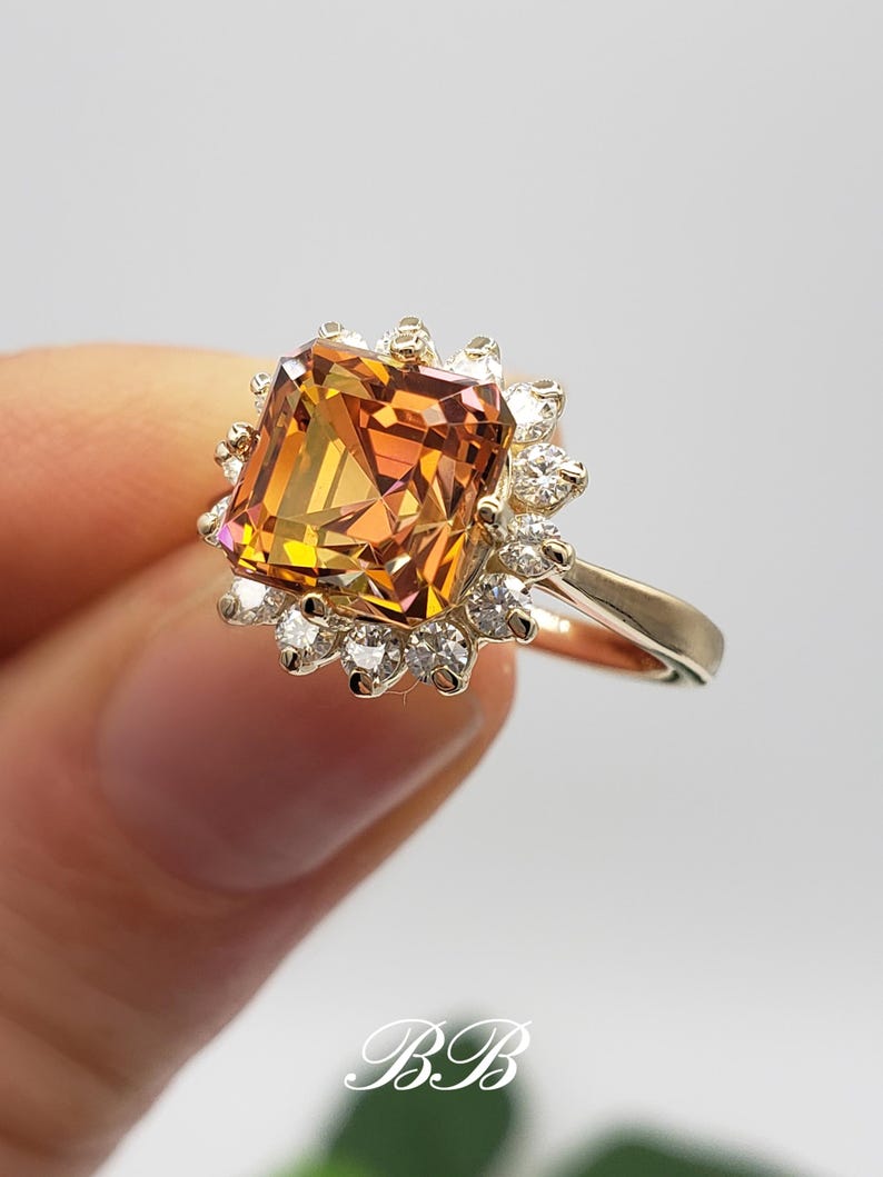 Pode incluir: Um anel de ouro com uma grande gema quadrada, de cor laranja e marrom, cercada por um halo de gemas brancas menores.