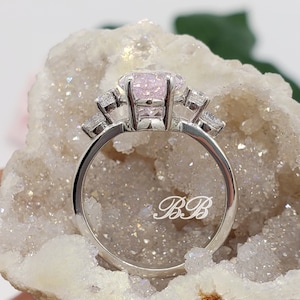 K&ouml;nnte beinhalten: Ein silberner Ring mit einem rosafarbenen Edelstein und kleineren klaren Edelsteinen auf der Ringschiene. Der Ring ist mit den Initialen "BB" graviert.