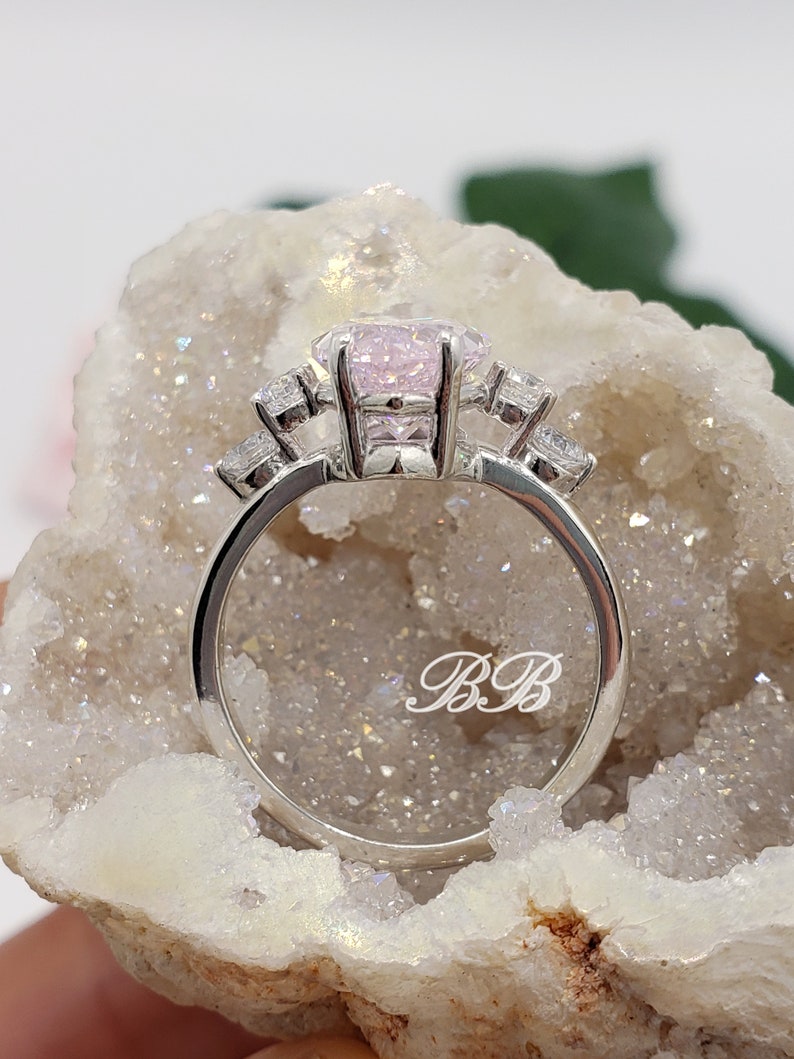 K&ouml;nnte beinhalten: Ein silberner Ring mit einem rosafarbenen Edelstein und kleineren klaren Edelsteinen auf der Ringschiene. Der Ring ist mit den Initialen "BB" graviert.