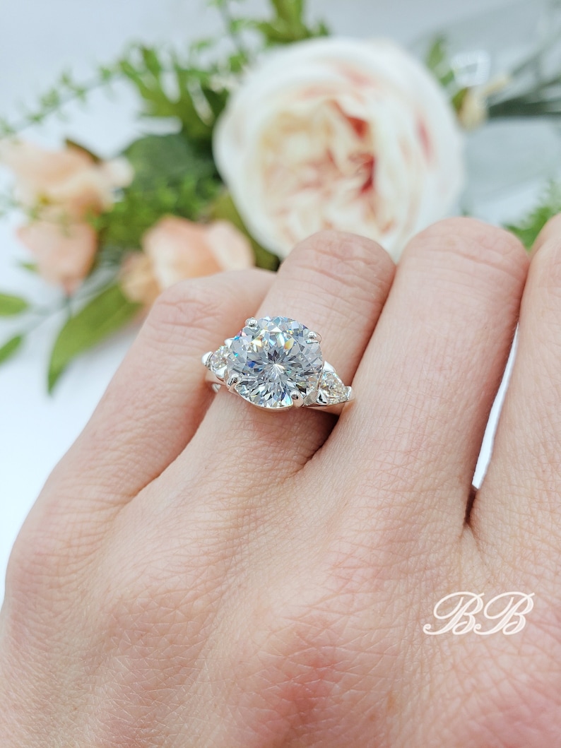 May include: A silver engagement ring with a large, round, clear gemstone as the centerpiece. Two smaller, triangular gemstones flank the main stone. The ring is set against a soft, blurred floral background.