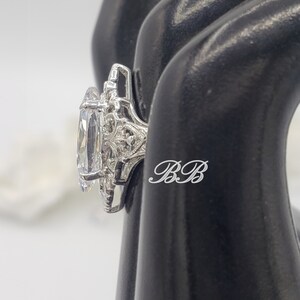Puede incluir: Un anillo de plata con una gran piedra preciosa ovalada transparente rodeada de piedras preciosas transparentes m&aacute;s peque&ntilde;as. El anillo est&aacute; colocado en un expositor de anillos negro.