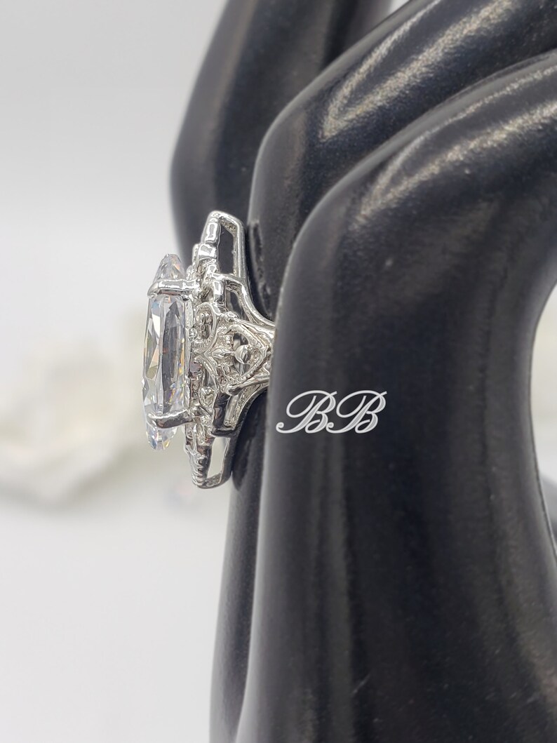 Puede incluir: Un anillo de plata con una gran piedra preciosa ovalada transparente rodeada de piedras preciosas transparentes m&aacute;s peque&ntilde;as. El anillo est&aacute; colocado en un expositor de anillos negro.