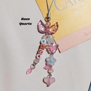 May include: A pink and gold beaded charm with a butterfly and angel wing design. The charm includes a star, flower, heart, and beads spelling out "SCC". The text "Rose Quartz" is also visible.