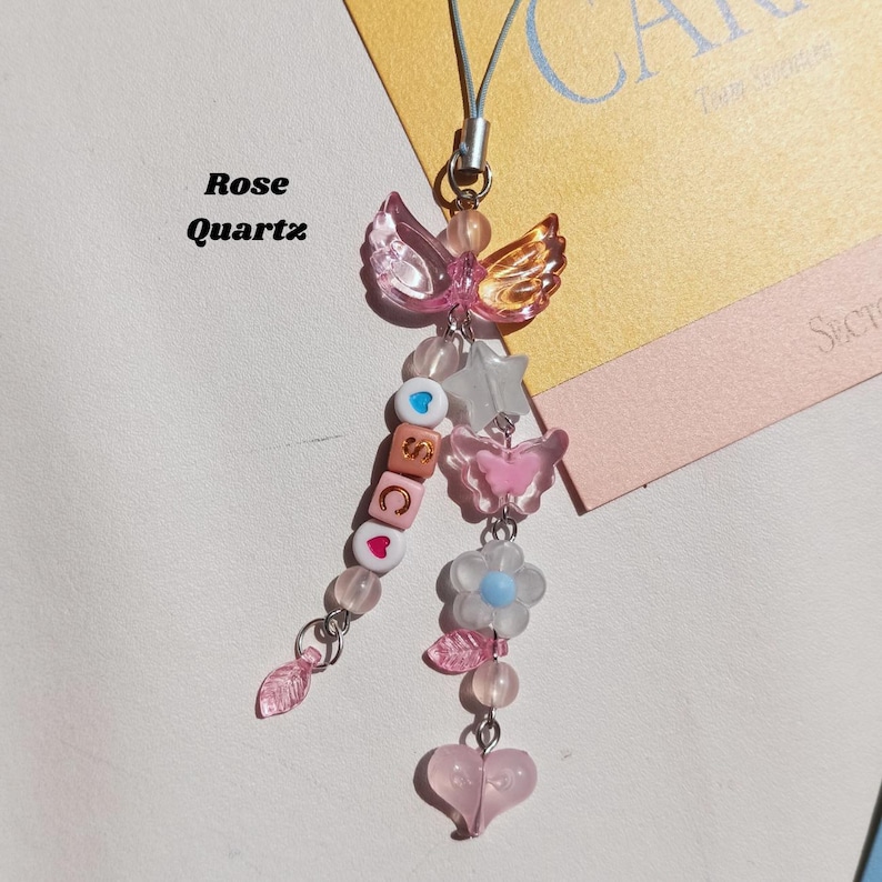 May include: A pink and gold beaded charm with a butterfly and angel wing design. The charm includes a star, flower, heart, and beads spelling out "SCC". The text "Rose Quartz" is also visible.