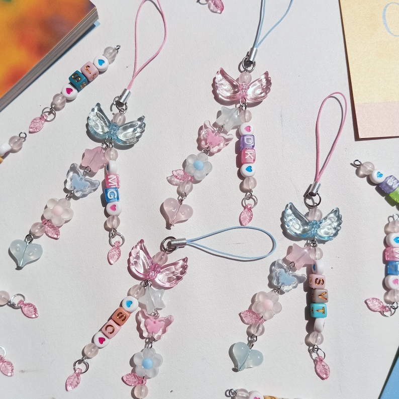 May include: A collection of handmade beaded phone charms in pastel colors. Each charm features a butterfly or angel wing design, along with heart and flower accents. Some charms include lettered beads spelling out initials or words. The charms are attached to a loop.