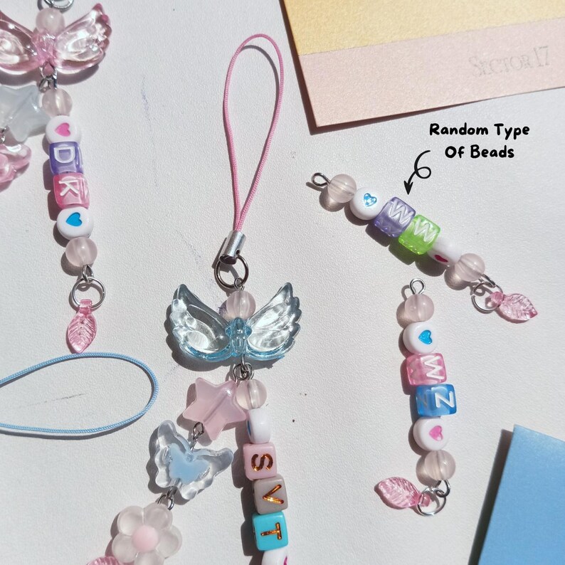May include: A collection of handmade beaded phone charms in pastel colors. The charms feature clear angel wings, star, and flower charms, along with letter beads spelling out words. The text "Random Type Of Beads" is also visible.
