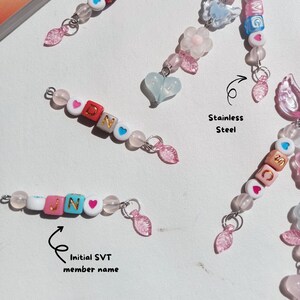 May include: Close-up of beaded keychains with the initials "Dino" and "JIN" spelled out with colorful letter beads. The keychains feature pink, white, and blue beads, with heart-shaped accents and pink leaf charms. The text "Stainless Steel" is also visible.