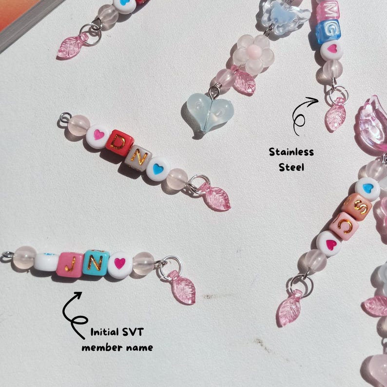 May include: Close-up of beaded keychains with the initials "Dino" and "JIN" spelled out with colorful letter beads. The keychains feature pink, white, and blue beads, with heart-shaped accents and pink leaf charms. The text "Stainless Steel" is also visible.