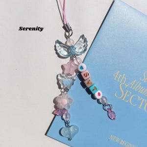 May include: A handmade beaded charm with a pink cord, featuring clear angel wings, a pink star, and a flower. The charm includes beads spelling "SVT" and a heart. The word "Serenity" is printed in black.