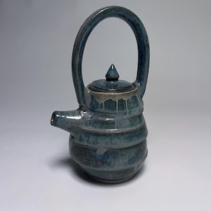 May include: A handmade ceramic teapot with a blue glaze. The teapot has a rounded body with horizontal ridges, a curved handle, a spout, and a lid with a pointed knob. The color is a mix of blues and greens, with some areas appearing lighter.