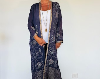 Könnte beinhalten: Eine lange, dunkelblaue, durchsichtige, bestickte Jacke mit goldenen Verzierungen und Pailletten. Die Jacke hat einen V-Ausschnitt und lange Ärmel. Das Model trägt ein weißes Oberteil und eine schwarze Hose.