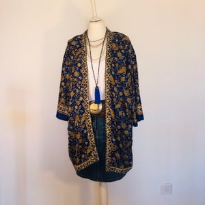 May include: A blue and gold floral print kimono jacket, white top, and denim skirt. The jacket has dark blue sleeve trim. A long beaded necklace with a blue tassel and a silver belt complete the outfit. The outfit is displayed on a mannequin.