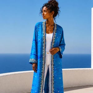 Pode incluir: Um quimono longo azul aberto na frente com um padrão ondulado subtil, bordados metálicos intrincados ao longo das lapelas e punhos, e pequenos adornos prateados por todo o tecido, usado sobre uma camisola sem mangas e calças brancas.