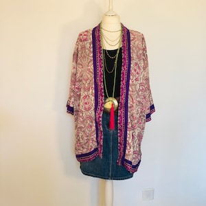 May include: A patterned kimono in cream, pink, and purple, displayed on a mannequin. The kimono features a floral design and is paired with a black top, denim skirt, and layered necklaces. A large pendant with a pink tassel adds a pop of color.