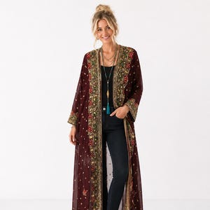 Könnte beinhalten: Ein bodenlanger, transparenter, burgunderroter Duster-Cardigan mit aufwendigen Goldstickereien, Pailletten und roten Blumenakzenten entlang der Vorderseite und an den Ärmelbündchen, getragen über einem schwarzen Top, einer dunkelblauen Jeans und goldenen Sandalen.