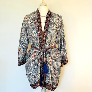 May include: A patterned kimono-style robe in cream, blue, and red paisley print. The robe has long sleeves, a tie belt, and a blue tassel. The trim is a dark red and black. The robe is displayed on a mannequin.