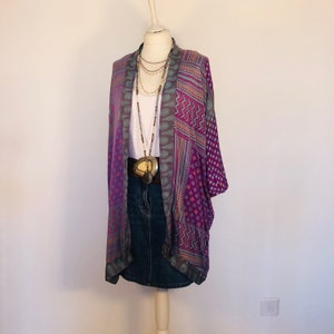 May include: A mannequin displays a layered outfit. A patterned purple and teal kimono is draped over a white tank top and denim skirt. A long necklace and a wide belt with a large pendant complete the look. The kimono features geometric and floral designs.