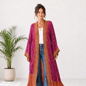 Puede incluir: Un kimono largo tipo duster abierto por delante en color magenta vibrante con un patrón abstracto morado oscuro. La prenda presenta mangas anchas y un borde ancho en contraste de color naranja quemado con detalles de bordado dorado.