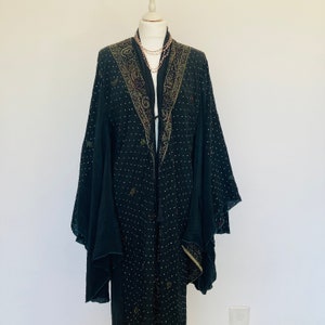 May include: A black, open-front kimono-style robe with intricate gold and bronze embroidery along the collar and cuffs. The robe is adorned with small, scattered white dots and hangs on a mannequin. A delicate necklace is draped on the mannequin.