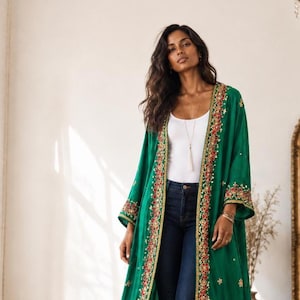 Pode incluir: Um casaco comprido verde-esmeralda aberto à frente, com bordados dourados intrincados e detalhes florais vermelhos ao longo das lapelas e dos punhos. A peça é usada sobre uma camisola branca simples e calças de ganga azul-escuras.