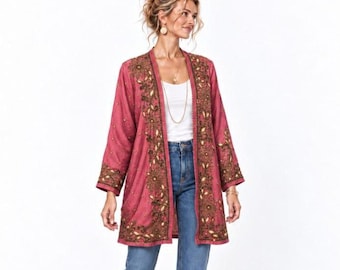 May include: A long-sleeved, open-front jacket in a dusty rose color featuring intricate dark embroidery and metallic accents along the lapels and cuffs, paired with a simple white tank top, blue denim jeans, and metallic gold flat shoes.