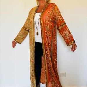 Può includere: Un lungo capo aperto con ricami dorati e arancioni. Il capo, simile a una veste, presenta un mix di motivi floreali e geometrici, con accenti di paillettes. Le maniche e i bordi sono rifiniti con dettagli dorati, creando un look fluido ed elegante.