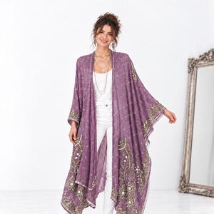Pode incluir: Um quimono longo roxo aberto na frente, com um padrão geométrico branco e uma borda floral verde-oliva na bainha e nas mangas. A peça é usada sobre uma regata branca simples e calças brancas, combinada com sandálias de salto alto.