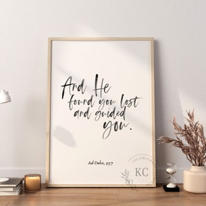 May include: A framed print with a white background and black calligraphy text that reads "And He found you lost and guided you." The text is attributed to Ad-Duba, 93:7. The print is framed in a light wood frame and is displayed on a white wall.