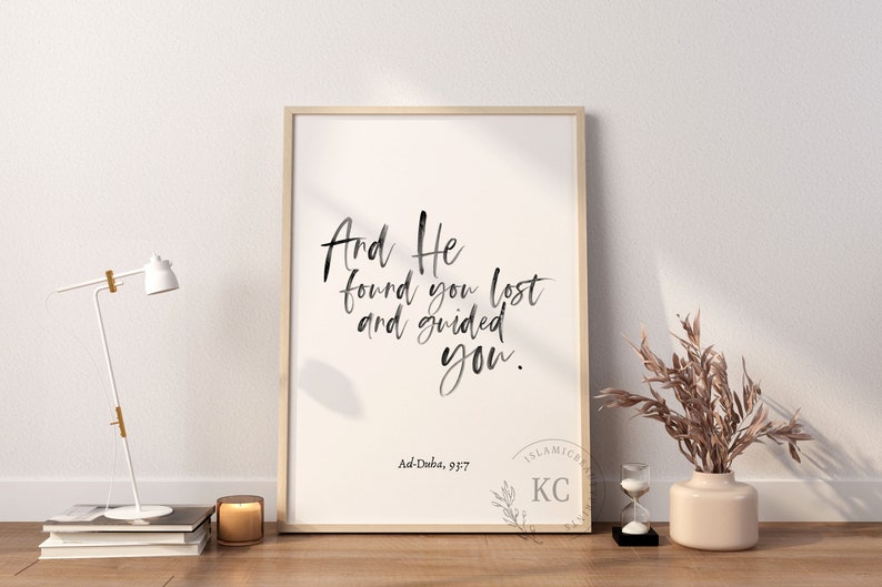 May include: A framed print with a white background and black calligraphy text that reads "And He found you lost and guided you." The text is attributed to Ad-Duba, 93:7. The print is framed in a light wood frame and is displayed on a white wall.