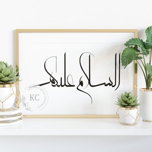 May include: A gold framed print with the Arabic calligraphy "Assalamu Alaikum" in black on a white background. The print is displayed on a white shelf with two potted succulents in gold geometric planters.