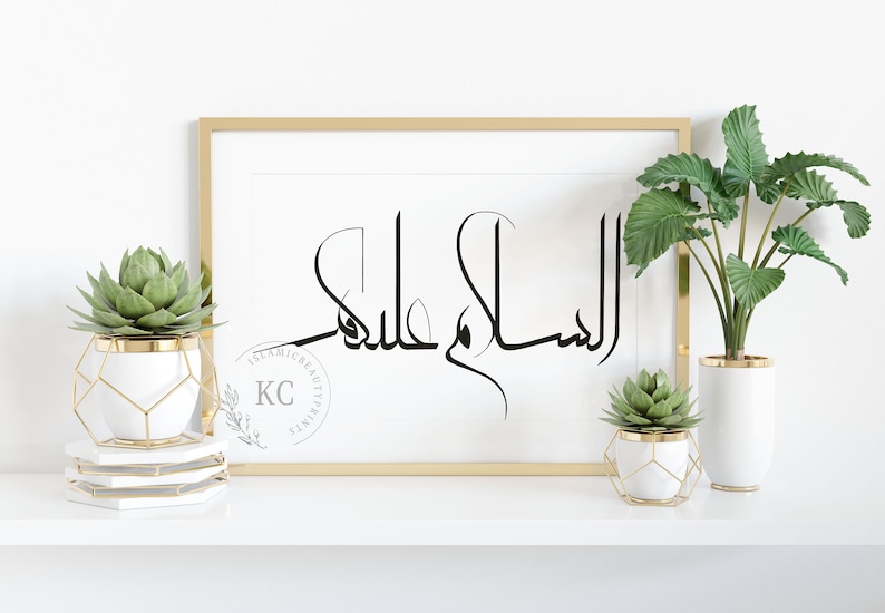 May include: A gold framed print with the Arabic calligraphy "Assalamu Alaikum" in black on a white background. The print is displayed on a white shelf with two potted succulents in gold geometric planters.
