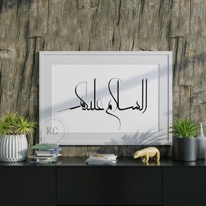 May include: A white framed print with black Arabic calligraphy that reads "Al Salam Alaykum" on a wooden background. The print is on a black shelf with a potted plant, books, and a small animal figurine.