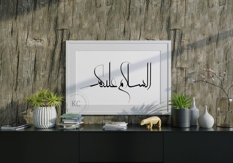 May include: A white framed print with black Arabic calligraphy that reads "Al Salam Alaykum" on a wooden background. The print is on a black shelf with a potted plant, books, and a small animal figurine.