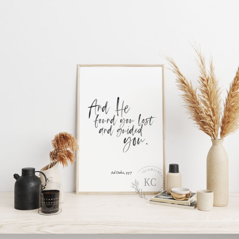 May include: A framed print with a black and white calligraphy quote that reads "And He found you lost and guided you." The quote is from the Islamic scripture Ad-Duha, 9:17. The print is on a white background and has a light wood frame.