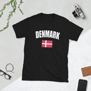 May include: A black short-sleeve t-shirt featuring the word DENMARK in white capital letters arched above a graphic of the red and white Danish flag. The shirt is made of a solid black fabric and displays the text and flag centered on the chest.